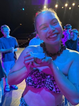 Smiling woman on stage holding an item, wearing patterned attire with a group in background.