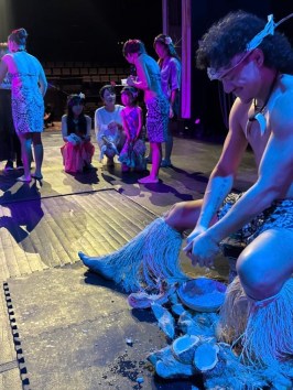 A person dressed in traditional attire working with natural materials on stage, surrounded by a group of people.