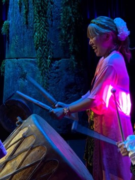 Women in colorful attire playing drums on stage with vibrant lighting.