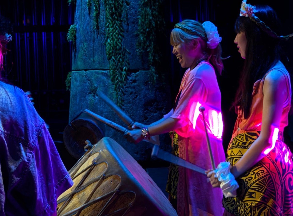Women in colorful attire playing drums on stage with vibrant lighting.