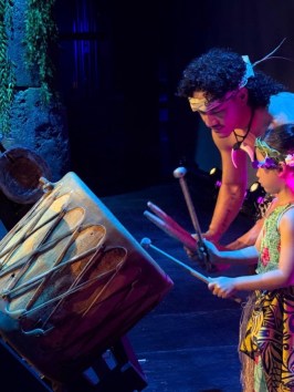 Two people in traditional attire play a large drum onstage.