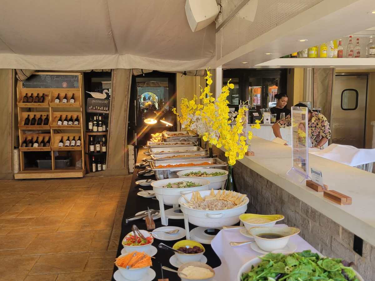 Buffet with salads, bowls, and platters under a canopy with bottles displayed on shelves.
