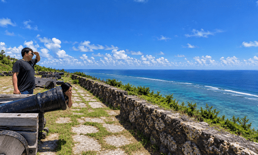 People looking out to sea beside old cannons, with blue sky and ocean in the background.