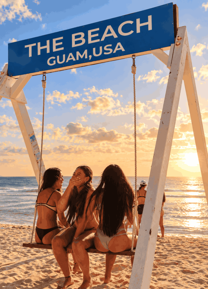 Three people on a swing at a beach with a sign overhead, during sunset.