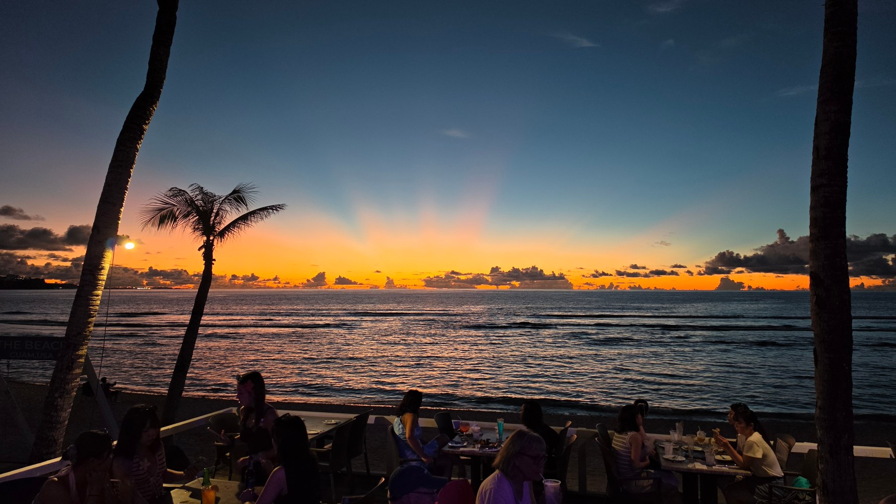 People dining at sunset by a tropical beach with palm trees.