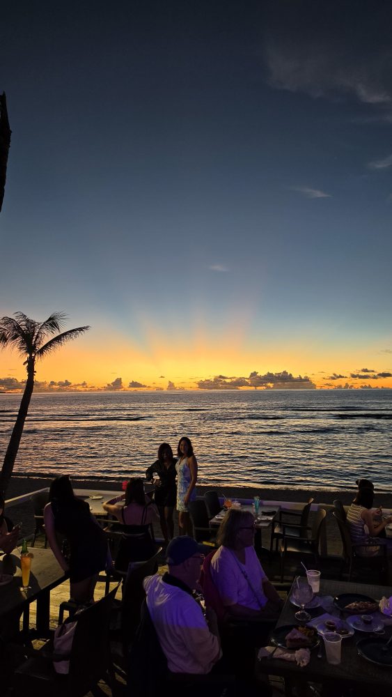 Sunset beach scene with people dining, palm tree, and ocean view.