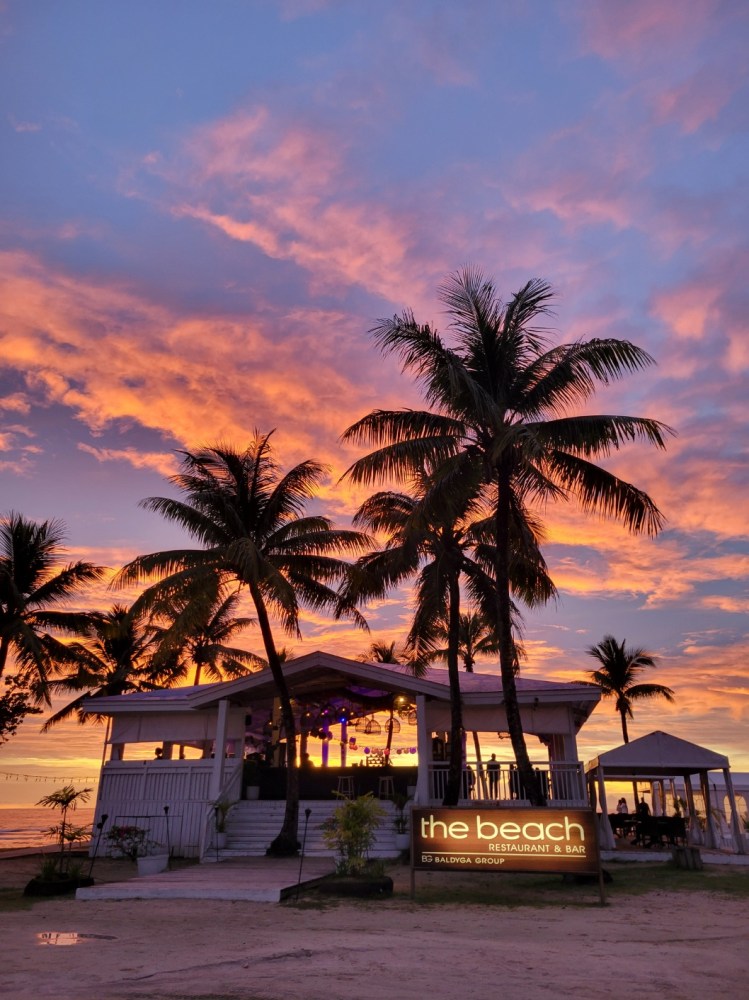 Beach restaurant with palm trees at sunset, vibrant orange and purple sky.